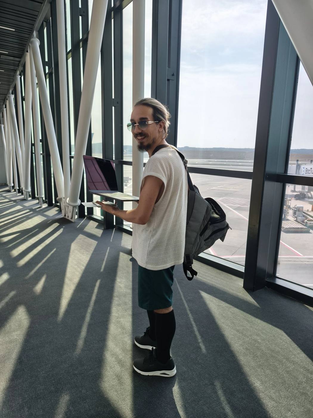 Me standing in an airport departure pier in low morning sun, an open laptop balanced on one hand and a backpack over one shoulder, with aircraft stands visible through the glass behind me