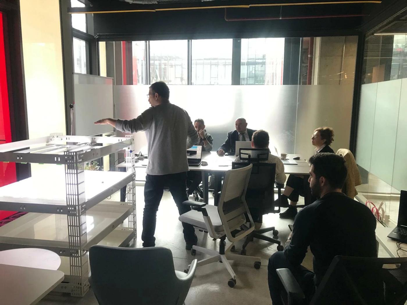 Seven people around a meeting table in a glass-walled office; a partly-assembled aluminium growing rack stands in the foreground, and one person is standing at a whiteboard with an arm out, describing a shelf spacing with his hands