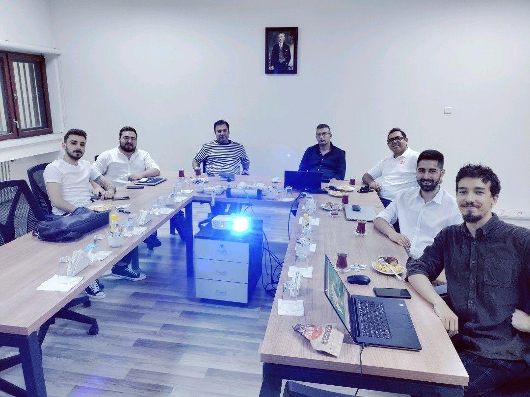 Eight people seated around a long conference table in a meeting room, a projector running in the middle of the table and laptops open along both sides